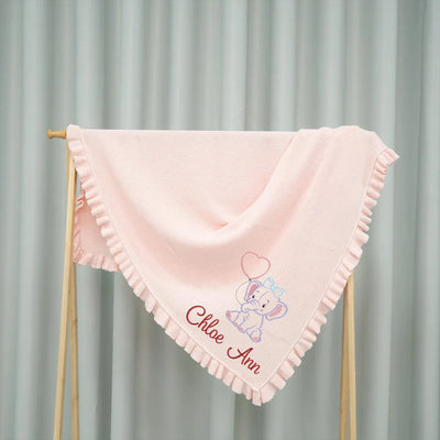 Pink personalized baby blanket with embroidered elephant design and text on a wooden stand against a gray curtain background