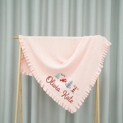 Pink personalized baby blanket with embroidered name and nautical designs on a stand against a gray curtain background