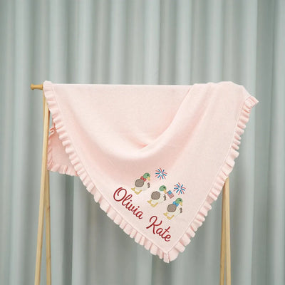 Pink personalized baby blanket with embroidered patriotic mallard duck trio design and text on a wooden stand against a gray curtain background