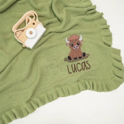 Sage green personalized baby blanket with a highland cow and 'Lucas' text, accompanied by a small camera toy.
